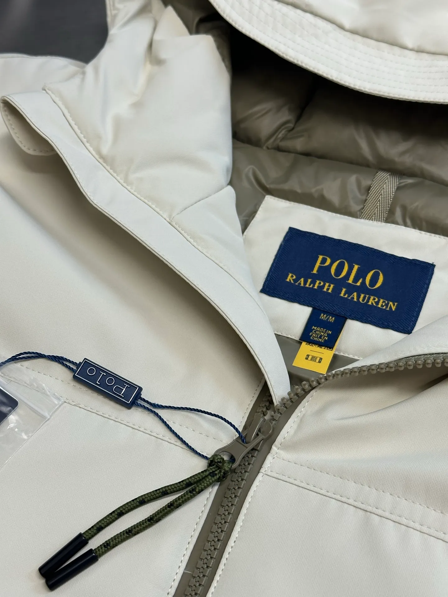 P970 Ralph Lauren logo hooded down jacket. The density of high-density nylon taffeta is about 210T-300T. The surface is smooth and slightly matte. It has excellent windproof and waterproof performance (with DWR water-repellent treatment). It has strong wrinkle resistance and is easy to care for. It is in line with the positioning of American casual "durable and practical".  The inner lining is made of 100% polyamide. The high-density and tight weaving process prevents fine lint from getting out of the gaps in the fabric. The smooth surface improves the putting on and taking off experience, and the excellent wear and tear resistance ensures damage caused by frequent use, while the light and soft texture does not affect the overall weight.  It uses 90 white duck down with a weight of 220g to ensure excellent thermal insulation performance. After strict screening and processing, the velvet is not easily drilled, ensuring durability. The classic jacket has a slightly loose fit, with slightly dropped shoulder lines (the shoulder width is 1-2cm wider than the standard fit). The length reaches the hips, and the sleeves are slightly tapered straight sleeves, taking into account freedom of movement and casual style.  The adjustable Velcro on the cuffs allows you to freely adjust the tightness according to the thickness of your wrist and personal preferences, effectively preventing cold wind from intruding, and improving the warmth performance. Two zippered pockets on the front are convenient for placing frequently used items, and the zipper design ensures that items are not easily lost. A built-in pocket on the left side of the chest provides a safe storage space for some small private items.  The drawstring design at the hem allows you to adjust the shape of the jacket according to your body shape and dressing needs. Whether it is loose and casual or slim and neat, you can easily control it.  Polo horse logo on the left chest: flat satin stitch embroidery (satin embroidery), using 100% polyester high color fastness embroidery thread (the color number is consistent with the brand's standard red color card), the stitch density is 12-15 stitches/cm, the outline of the horse logo is clear, and the details of the rider and horse are burr-free. Precise positioning through the template (12cm from the bottom of the collar and 8cm from the placket) ensures symmetry and centering.  size：s m l xl